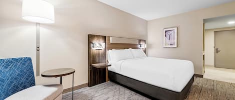 A hotel room with a bed, a small round table, a blue chair, a lamp, and a framed picture on the wall.
