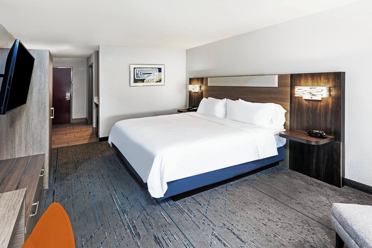 A modern hotel room with a large bed, a flat-screen TV, a wooden headboard, and a small table with a lamp.