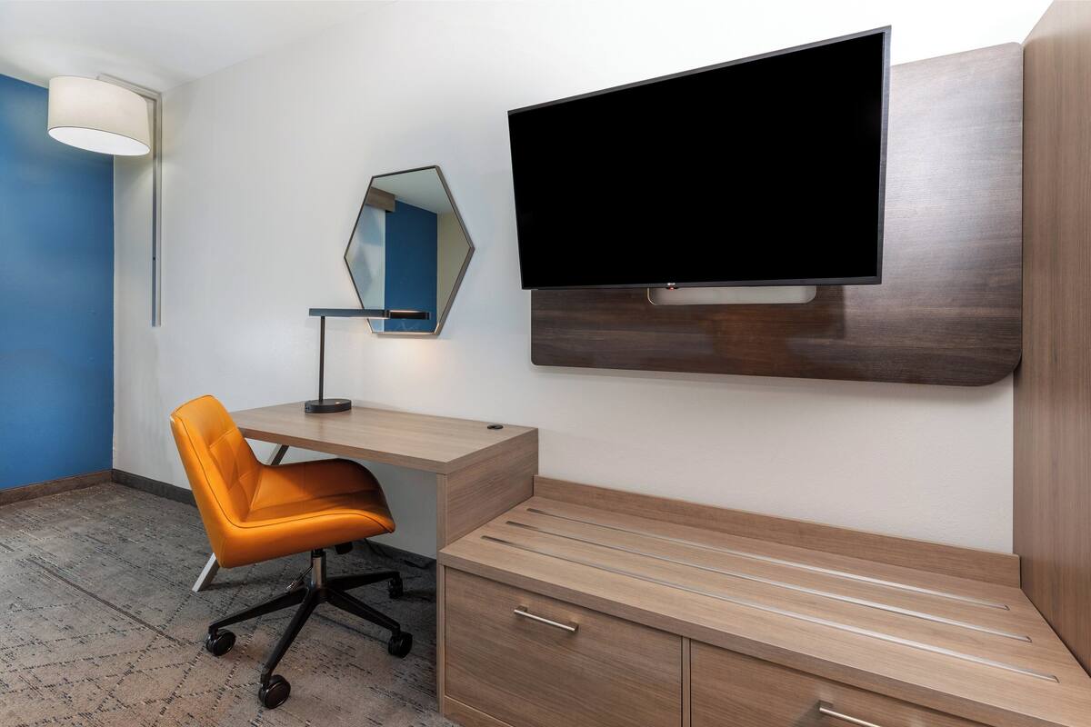 A modern hotel room with a wooden desk, an orange office chair, a flat-screen TV, a mirror, and a lamp.
