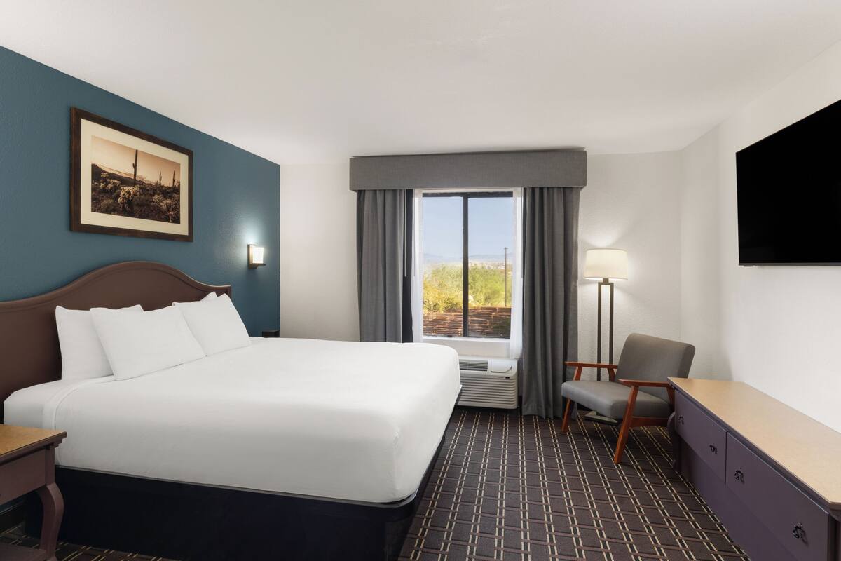 A hotel room with a bed, a desk, a chair, a window with curtains, a wall-mounted TV, and a framed picture.