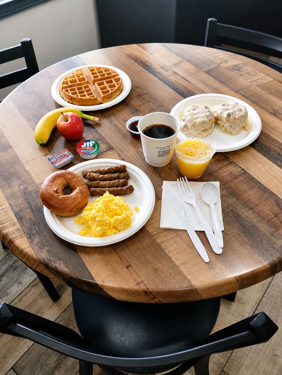 A breakfast table with waffles, sausages, scrambled eggs, a donut, a banana, an apple, a juice glass, and a coffee cup.