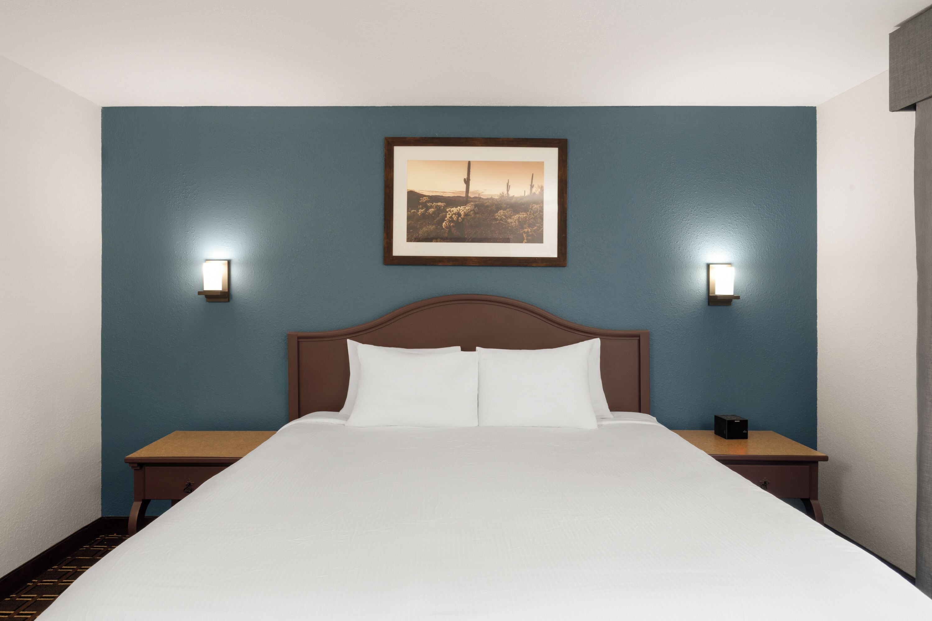 A hotel room with a bed, two bedside tables, a nightstand, a framed picture on the wall, and two wall-mounted lights.