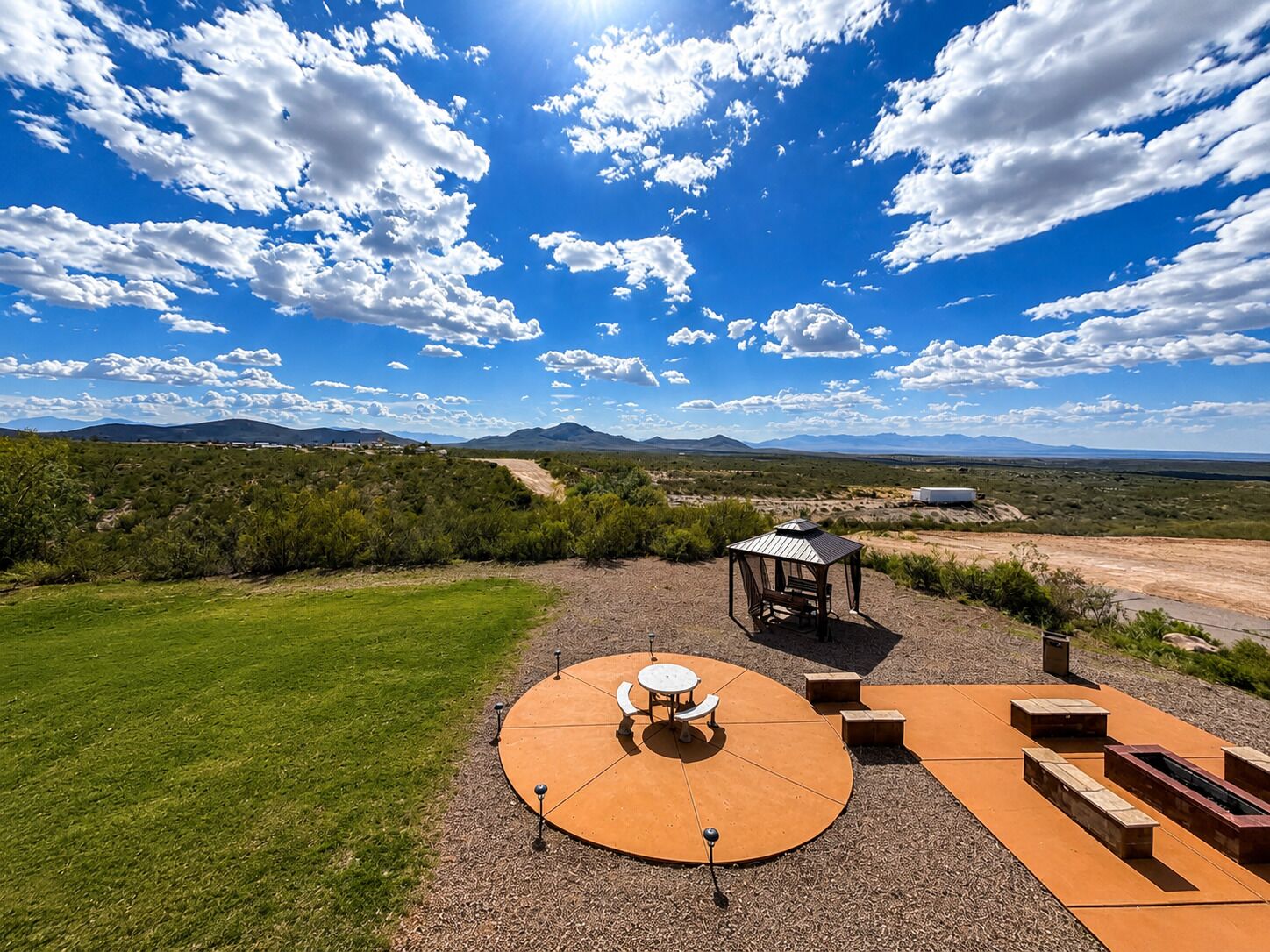 Eine Terrasse im Freien mit einer runden Sitzecke, einem kleinen Pavillon und Blick auf entfernte Berge unter klarem Himmel.