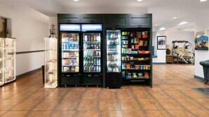 Un vestíbulo con una zona de máquinas expendedoras, que incluye un refrigerador lleno de bebidas y aperitivos, y un estante con diversos productos envasados.