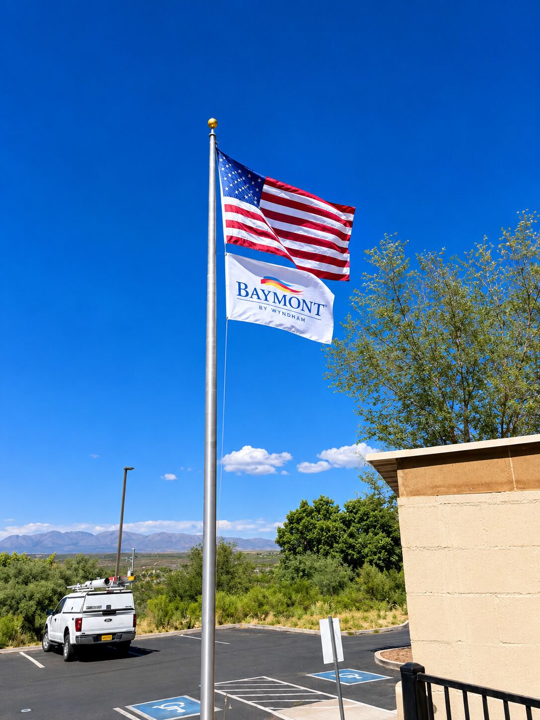 Un mât sur lequel flotte le drapeau américain, une enseigne Baymont By Wyndham, un parking avec un pick-up blanc et un ciel bleu dégagé.