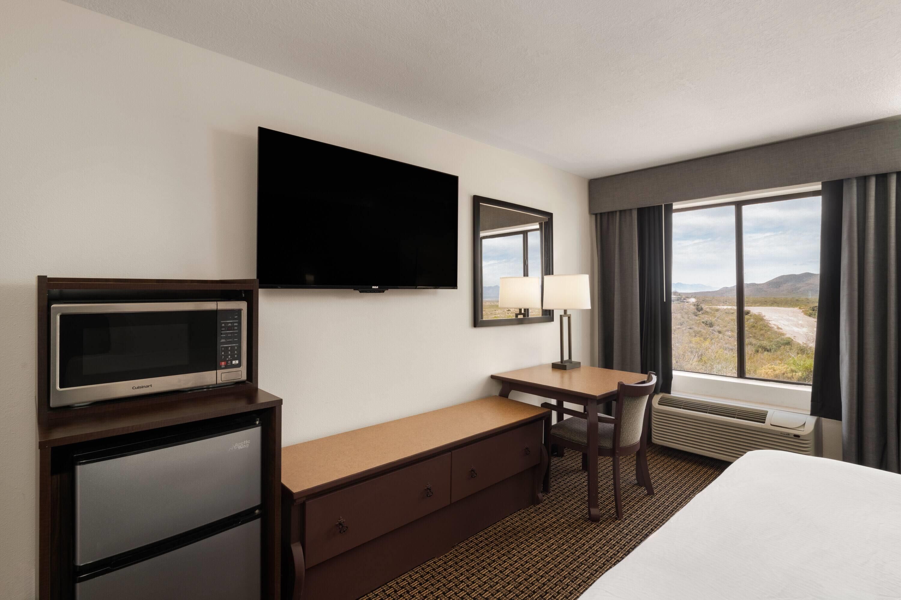 Habitación de hotel con cama, escritorio, silla, televisor de pantalla plana, microondas, refrigerador pequeño, ventana con vista al desierto y lámpara.