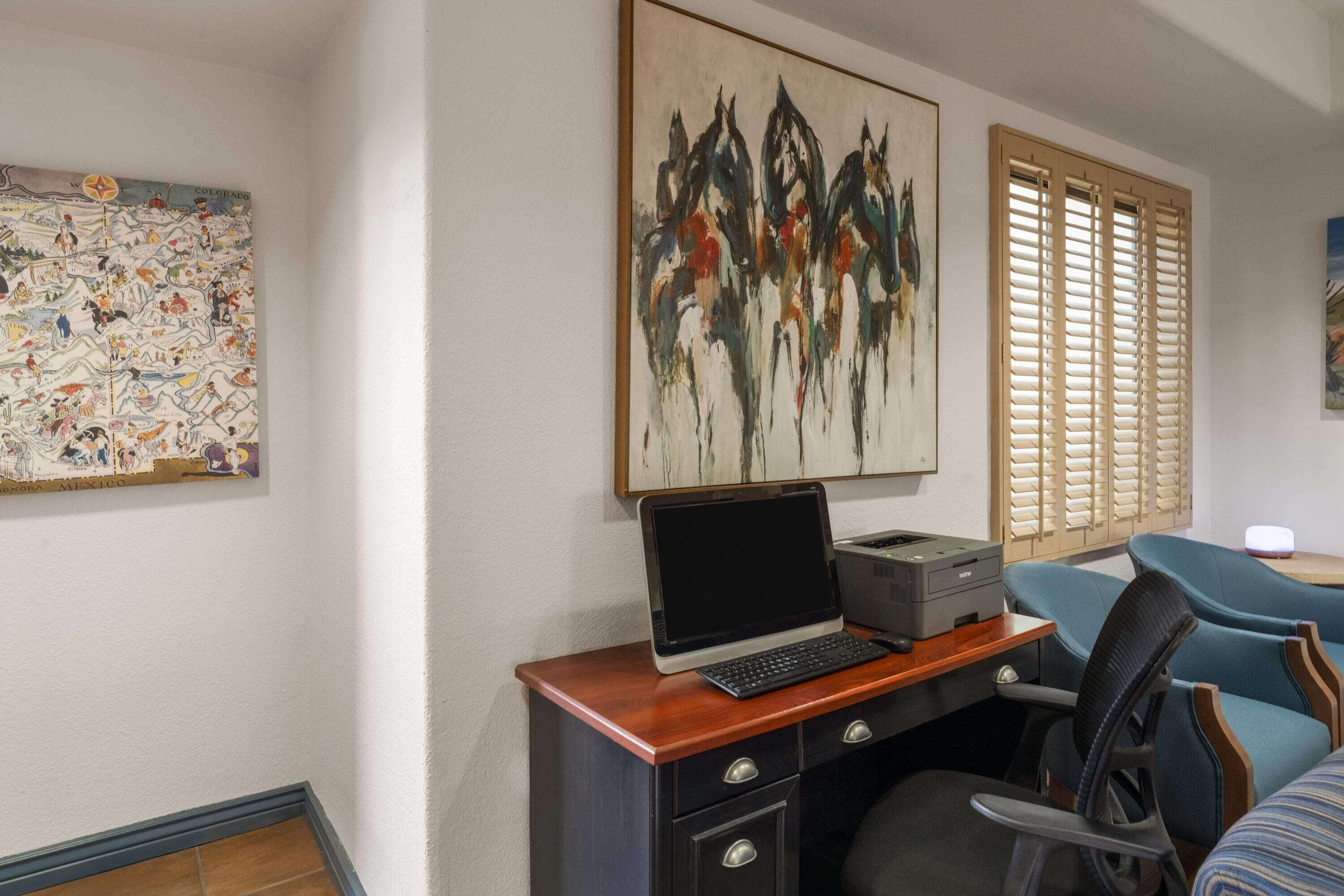 A room with a desk, a laptop, a printer, and a chair. There are two windows with blinds, and a painting on the wall.
