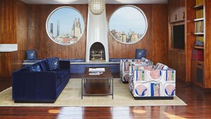Una sala de estar con chimenea, dos ventanas circulares con vistas a la ciudad, un sofá azul, un sofá estampado y una estantería.