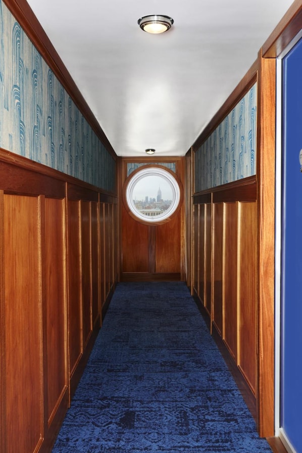 A hallway with wooden paneling, a blue carpet, and a round window with a cityscape view.