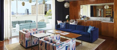 A modern living room with a blue sofa, patterned ottomans, and a wooden coffee table. A city view is visible through large windows, and a kitchen area is visible in the background.