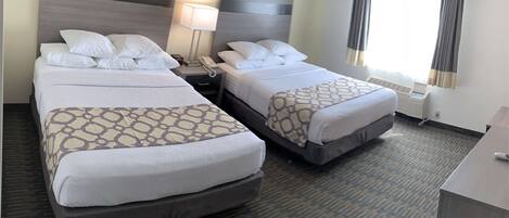 A hotel room with two beds, a bedside table with a lamp, a window with curtains, and a carpeted floor.