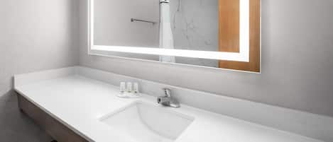 Un baño moderno con un lavabo blanco, un espejo grande y una ducha que se ve en el reflejo.