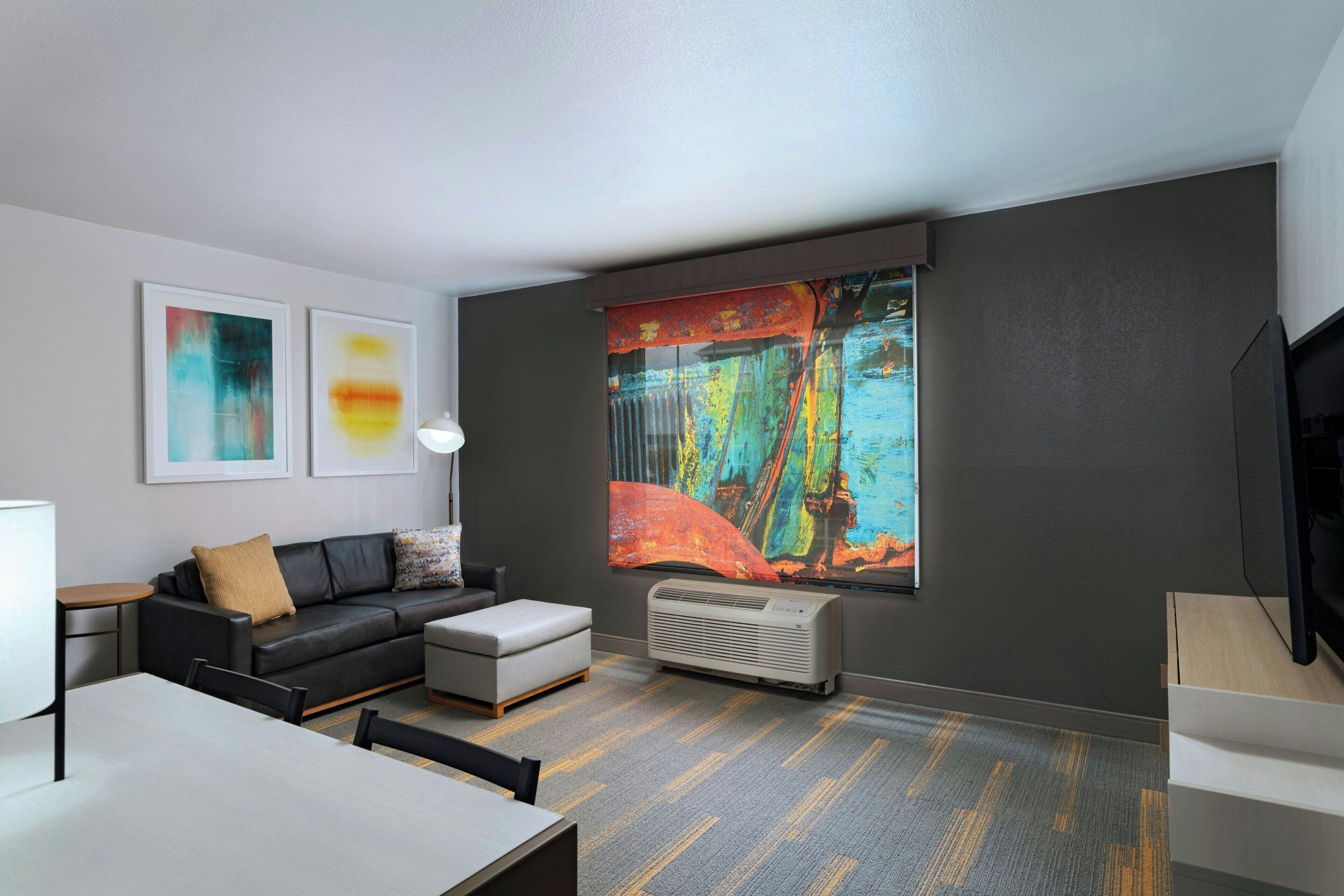 A modern living room with a black leather sofa, a gray ottoman, a dining table, and a large abstract painting on the wall.