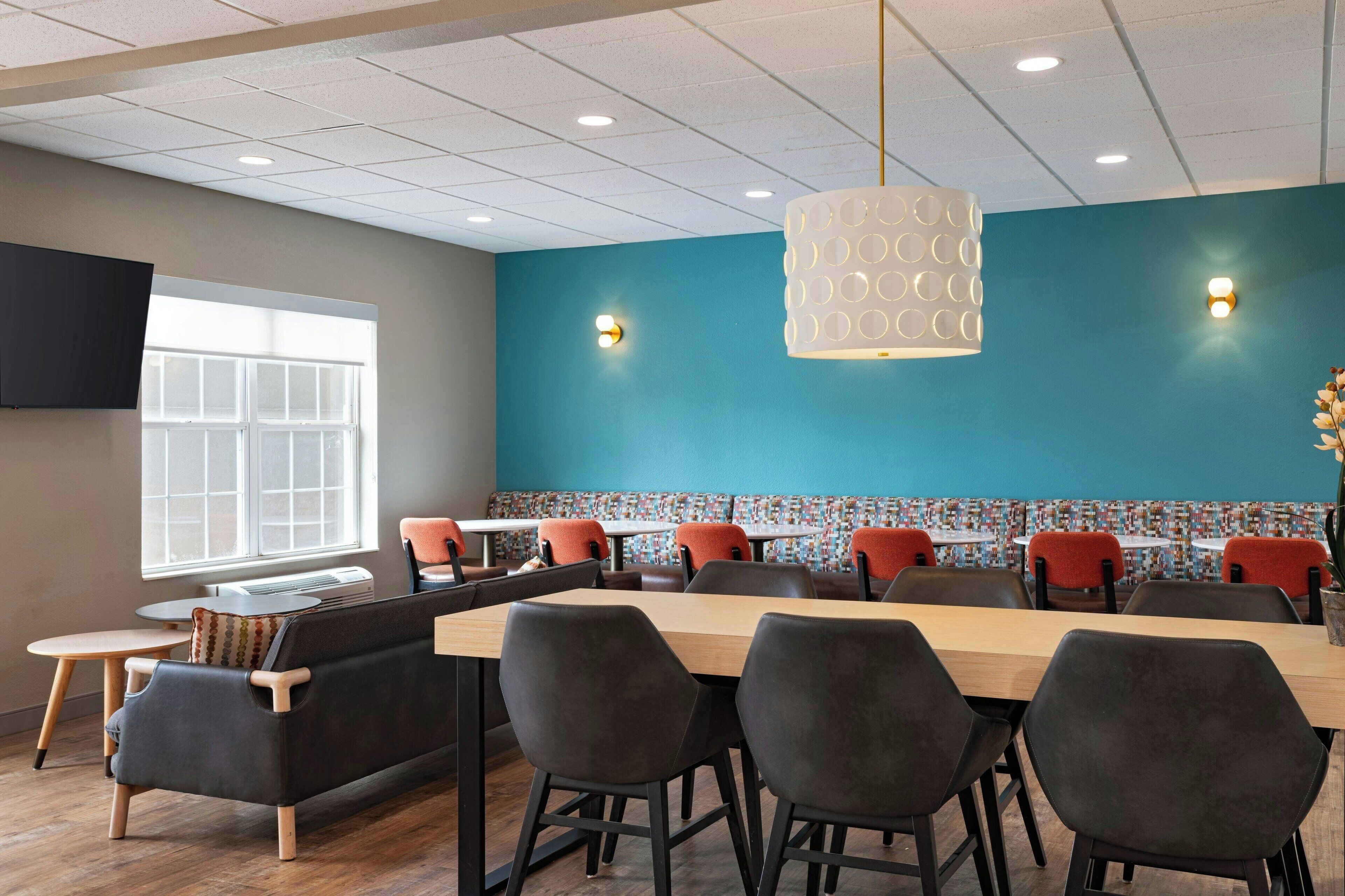A modern dining area with a long table, multiple chairs, and a sofa. There are two wall-mounted lights and a ceiling light fixture.