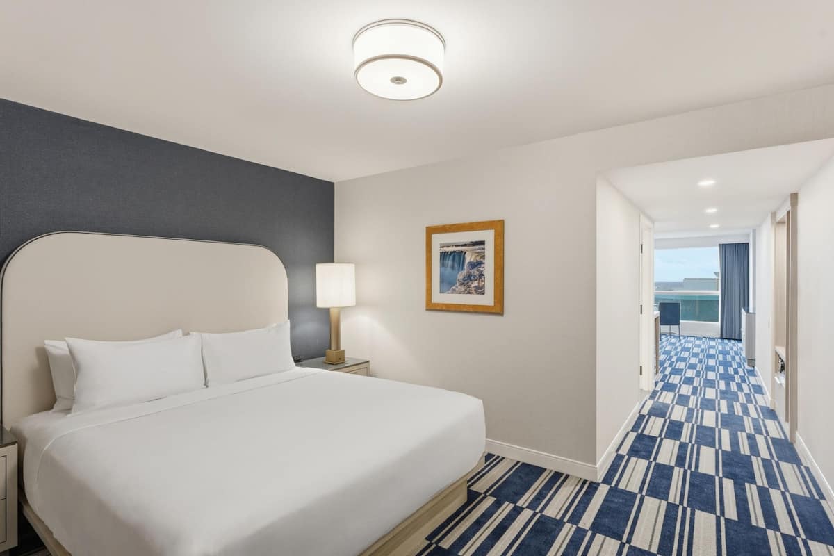 A hotel room with a large bed, a nightstand, a lamp, a framed picture on the wall, and a hallway with a patterned carpet.