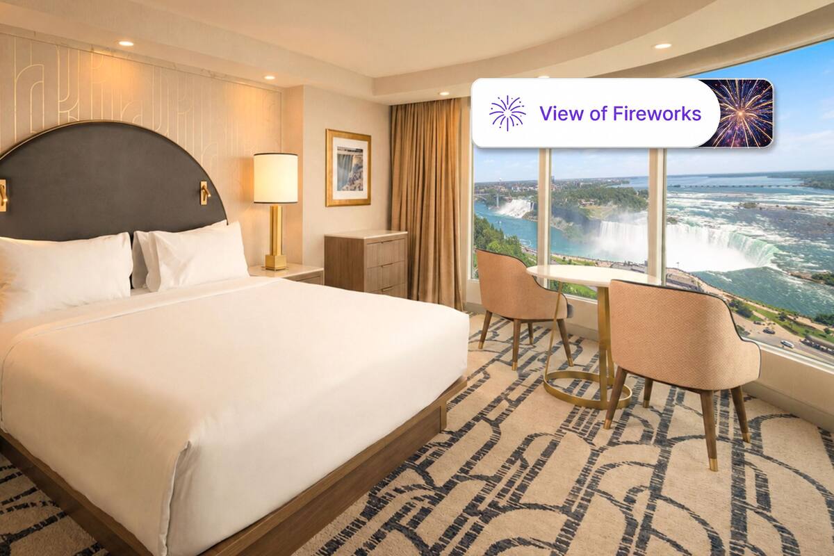 A hotel room with a large bed, two bedside tables with lamps, a desk with chairs, and a view of fireworks.