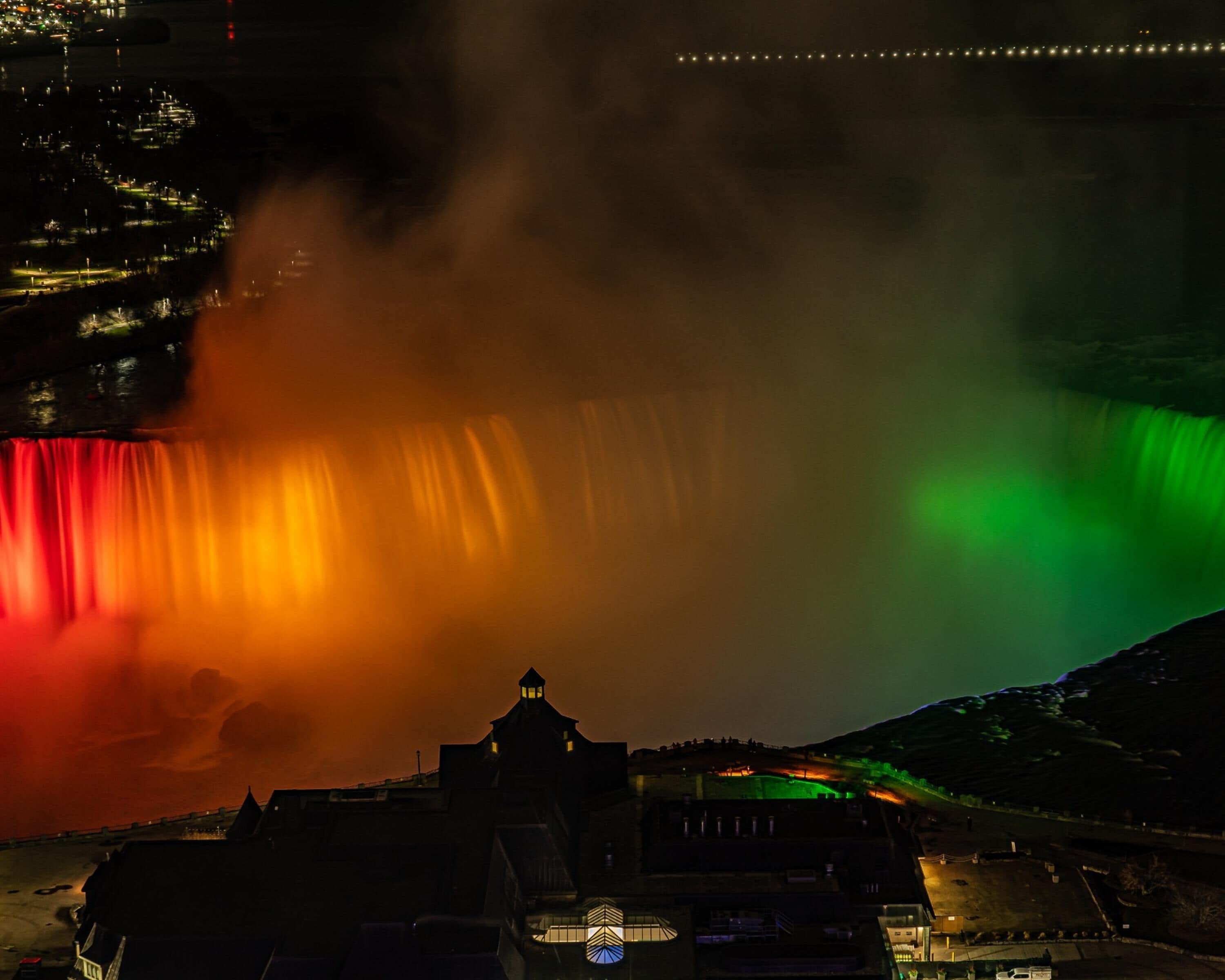 Een waterval met een meerkleurige lichtshow, een opvallend gebouw met een koepel en een heuvelachtig landschap op de achtergrond.