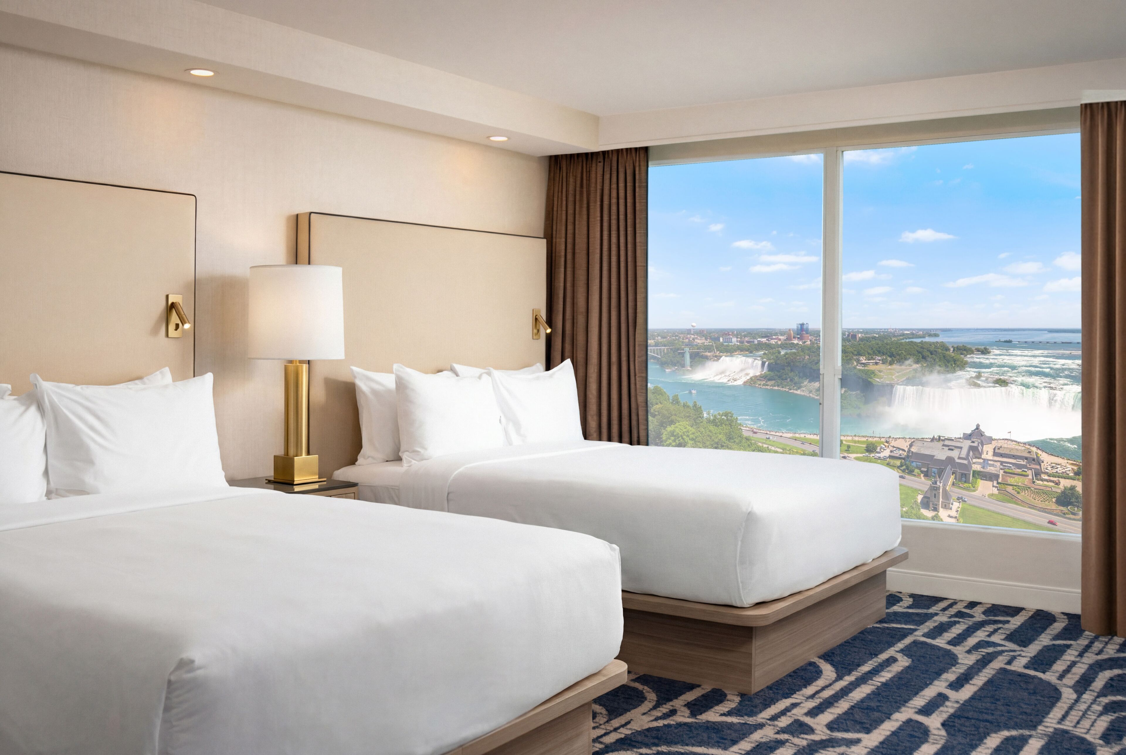 Ein Hotelzimmer mit zwei Betten, einem großen Fenster mit Blick auf eine Stadt und ein Gewässer, sowie einem blauen, gemusterten Teppich.