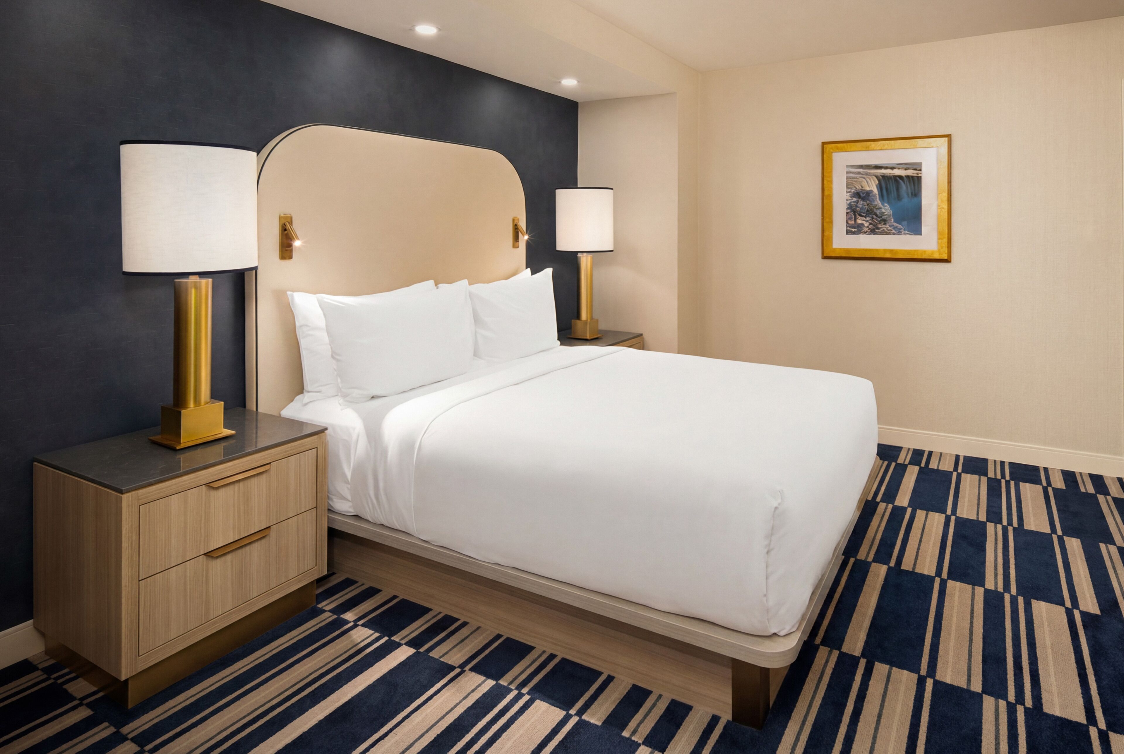 Una habitación de hotel con una cama, dos mesitas de noche con lámparas, una mesita con cajones, una alfombra estampada y un cuadro en la pared.