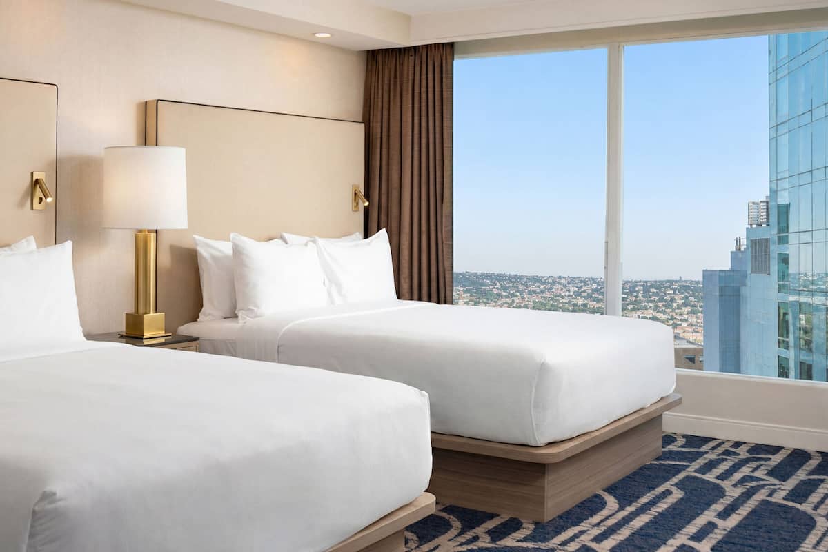 A hotel room with two beds, a large window with a city view, and a modern lamp.
