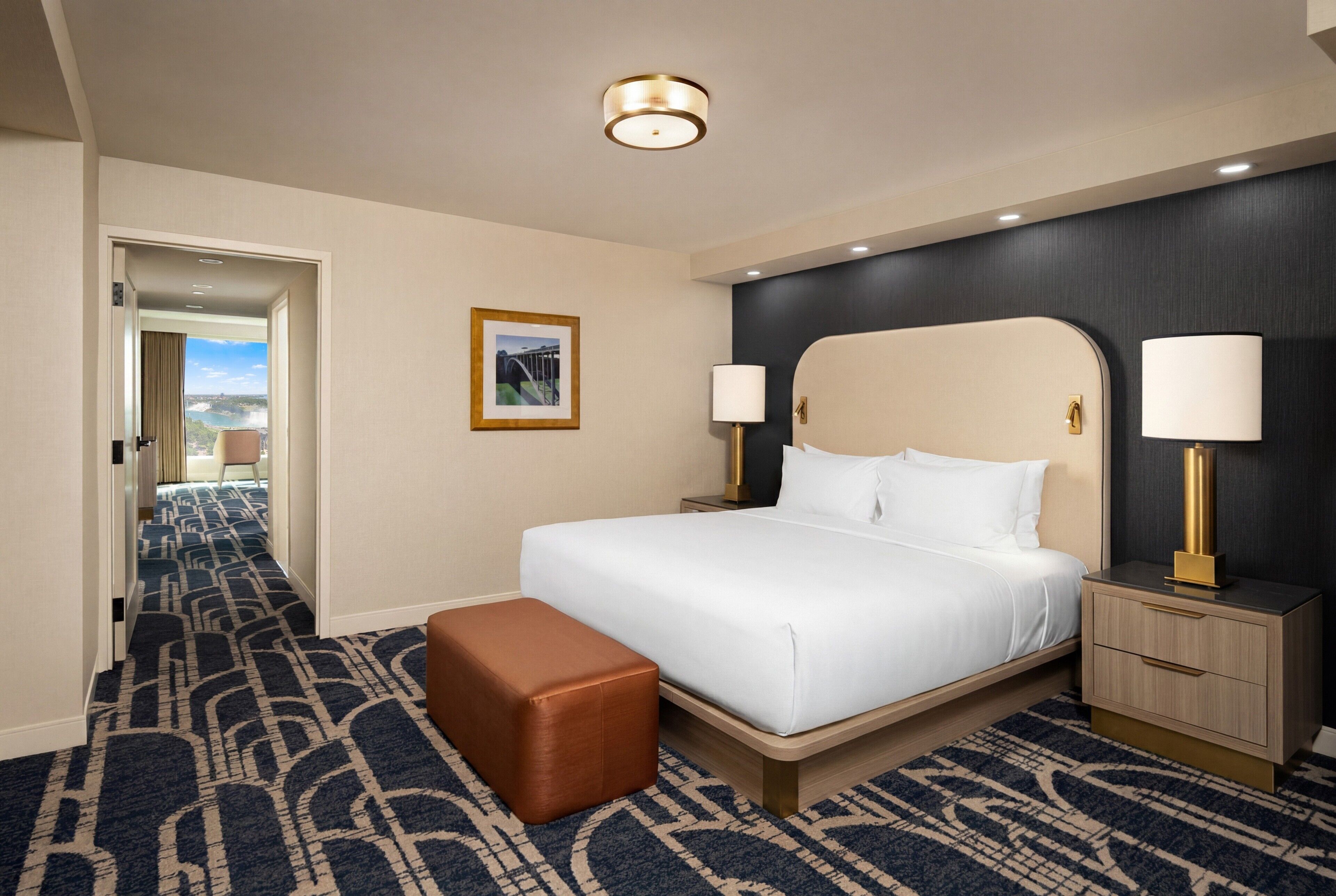 A hotel room with a large bed, a nightstand, a lamp, a framed picture on the wall, and a view through an open door.