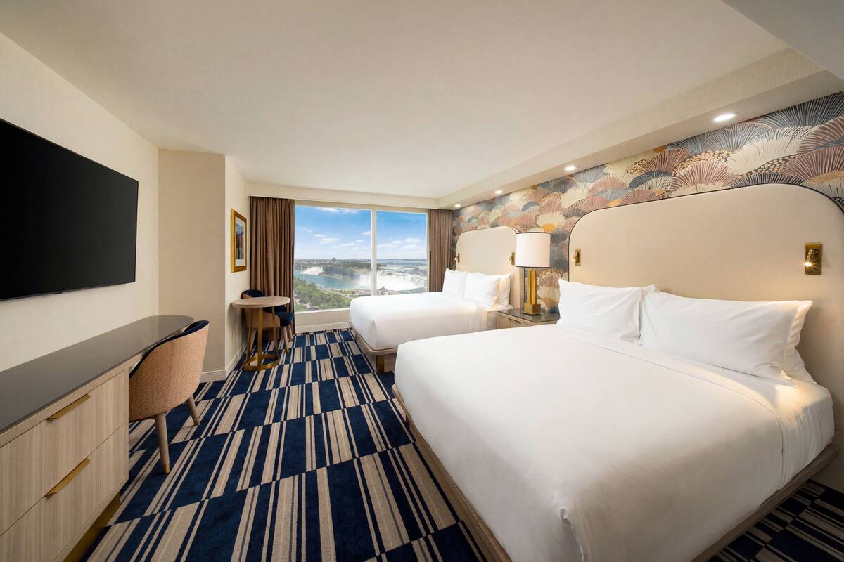 A hotel room with two beds, a desk, a TV, and a large window with a view.