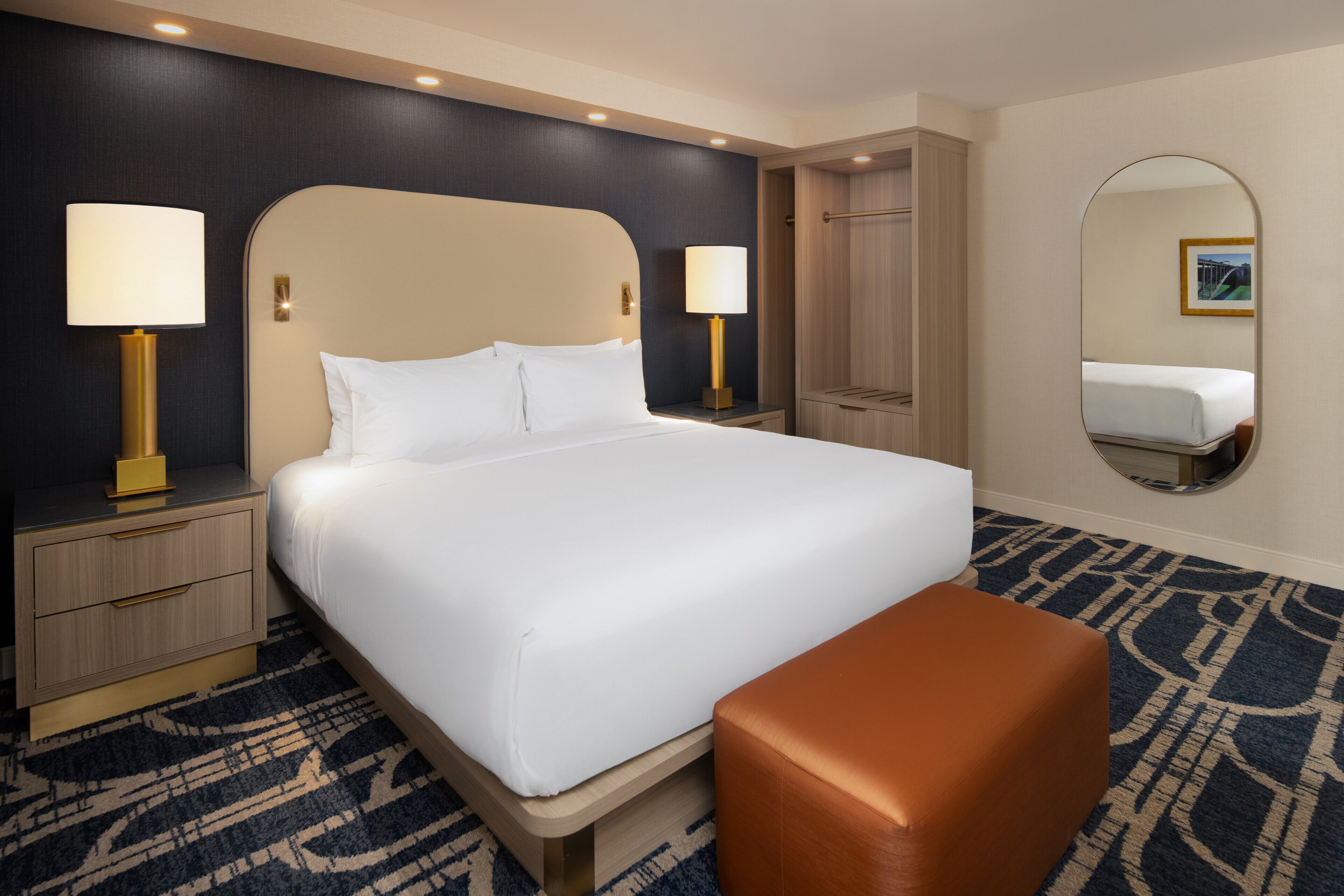 A hotel room with a large bed, two bedside tables with lamps, a wooden wardrobe, a round mirror, and a patterned carpet.