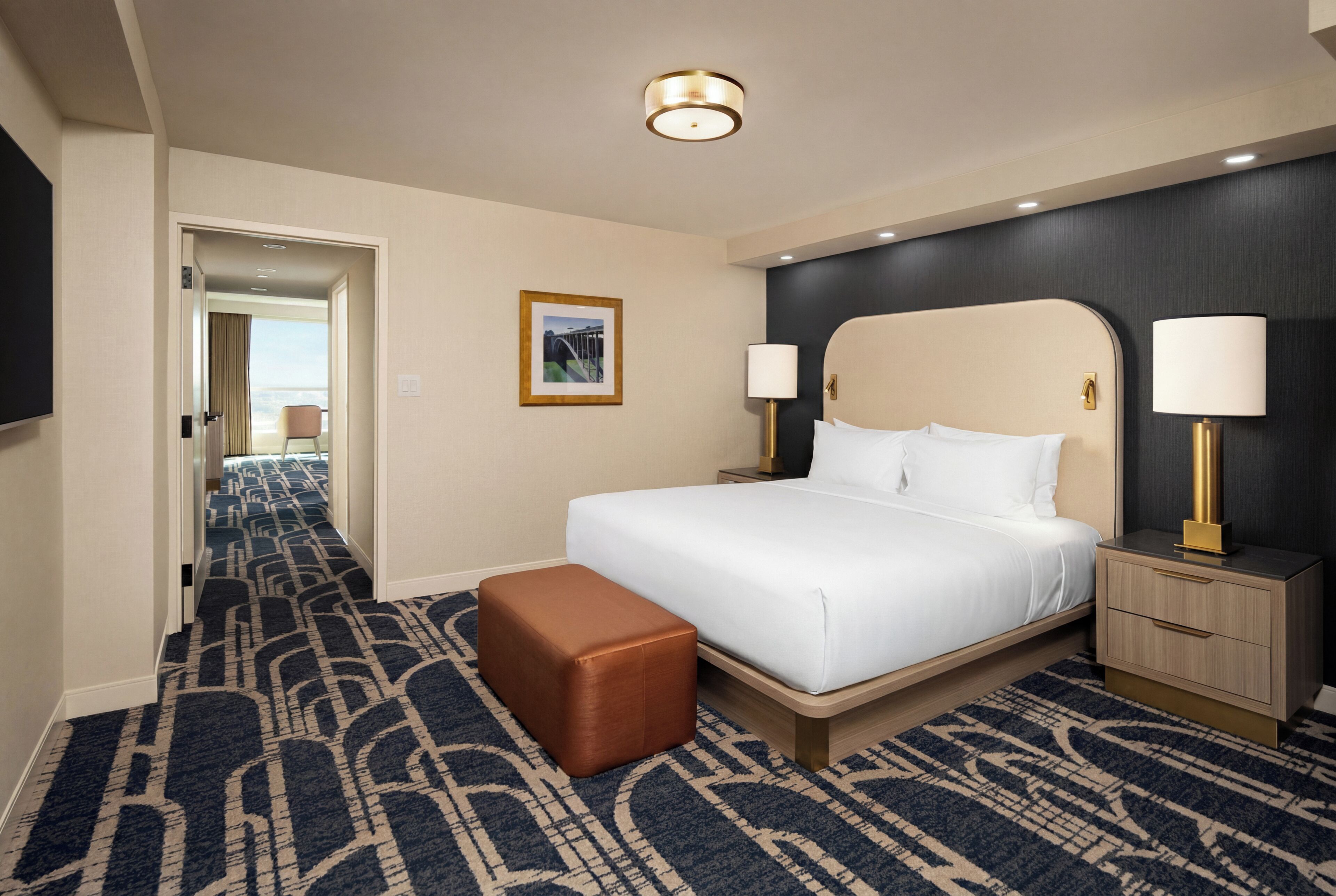 A hotel room with a large bed, a nightstand, a lamp, a picture on the wall, and a carpeted hallway.