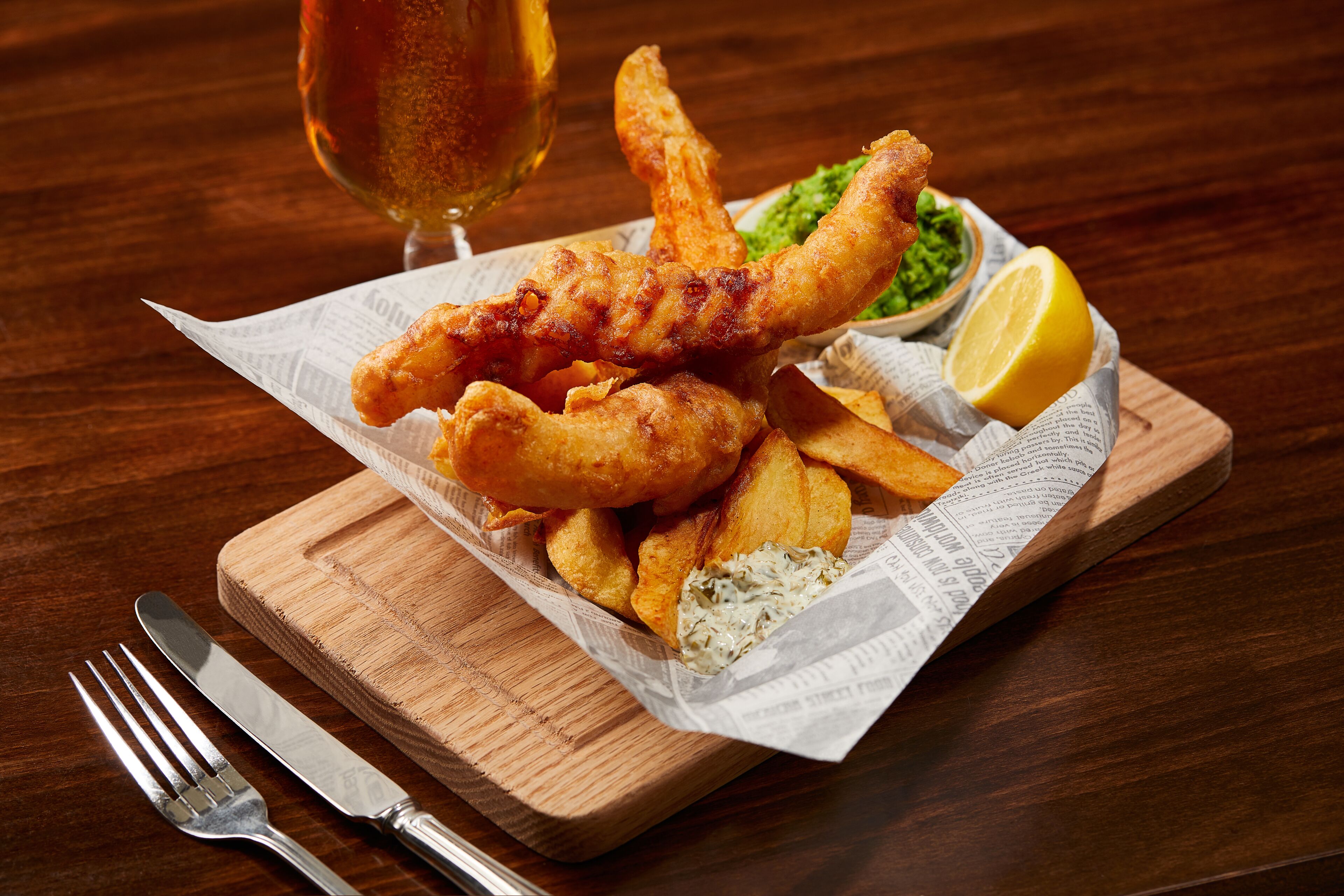 A meal of fish and chips on a wooden board with a newspaper underneath, accompanied by a fork and knife, and a glass of beer.