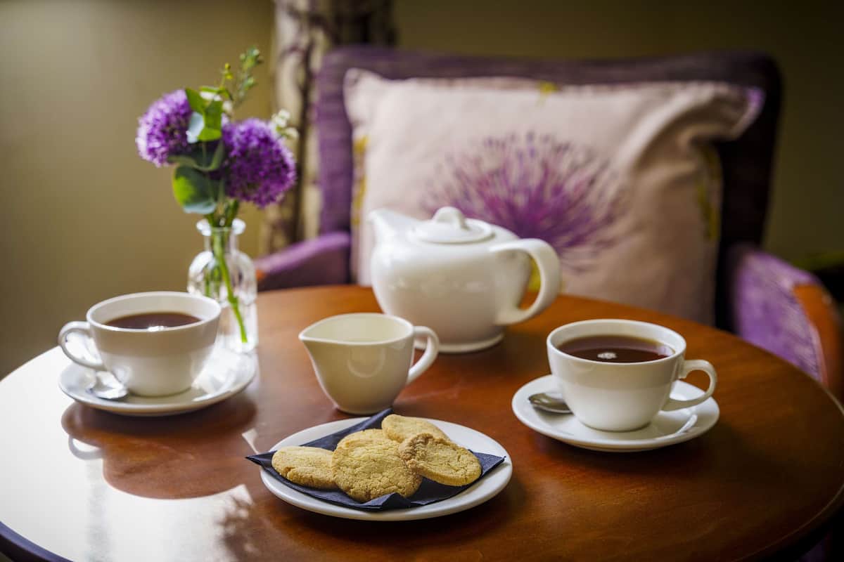 A round wooden table with two white cups of coffee, a white teapot, and a plate with cookies.