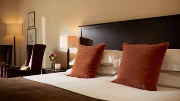 A hotel room with a large bed, two pillows, a nightstand with a lamp, a chair, and framed pictures on the wall.