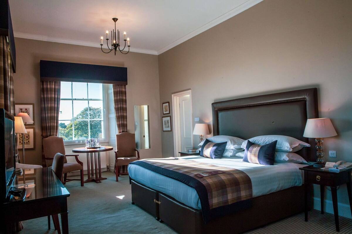 A hotel room with a large bed, a desk, a chair, a round table, and a chandelier.