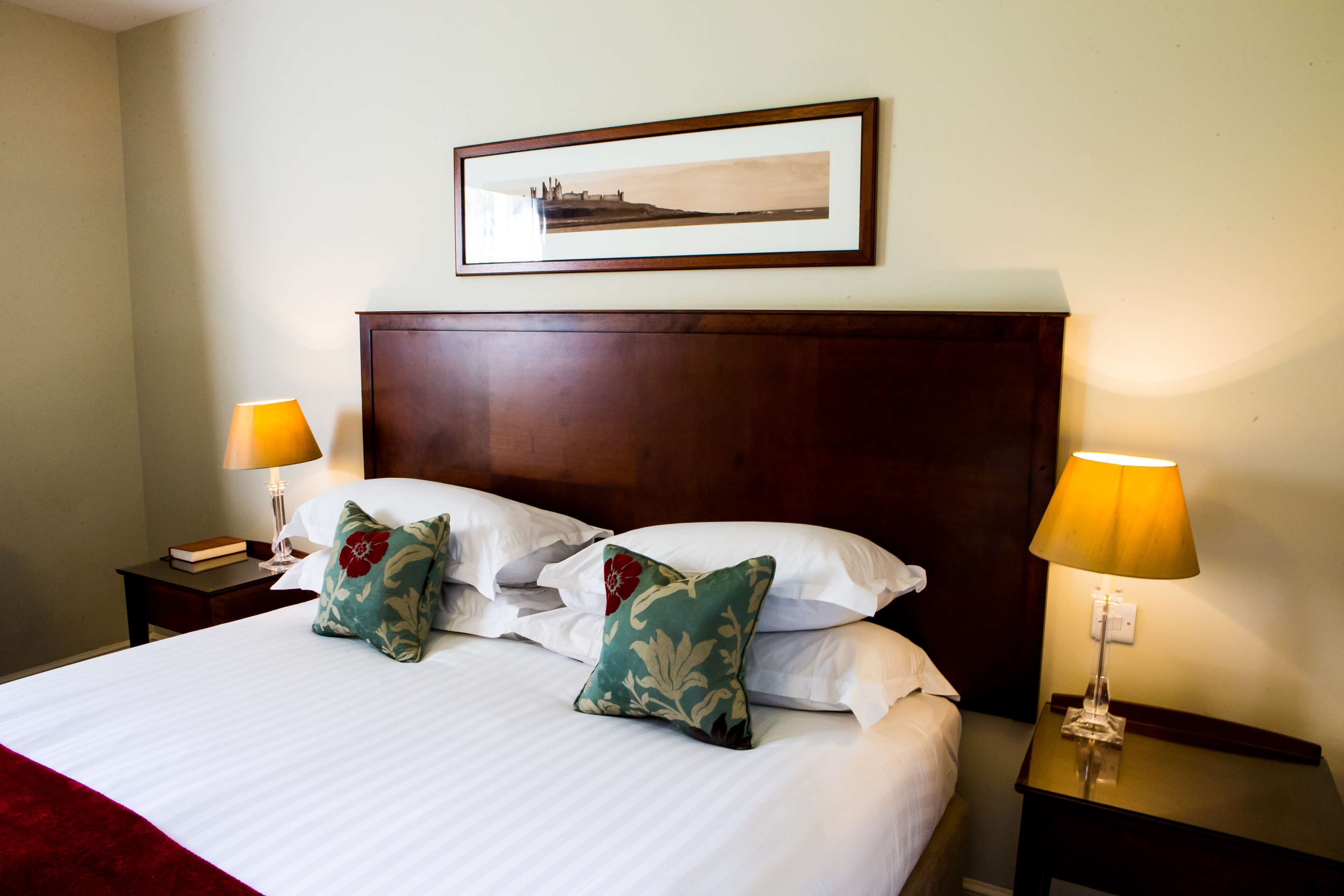 A neatly made bed with white linens, two decorative pillows, a framed picture above the bed, and two bedside lamps on wooden nightstands.