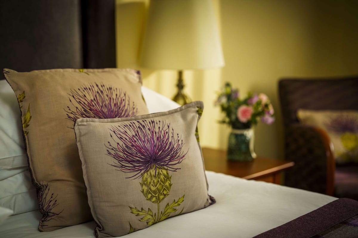 A bed with two decorative pillows featuring a floral design, a bedside table with a vase of flowers, and a lamp.
