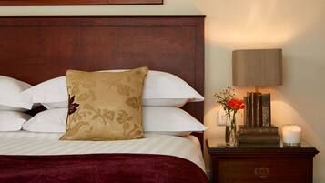 A neatly made bed with white and beige pillows, a red throw blanket, and a wooden headboard. A bedside table with a lamp, a vase with flowers, and a candle is visible.