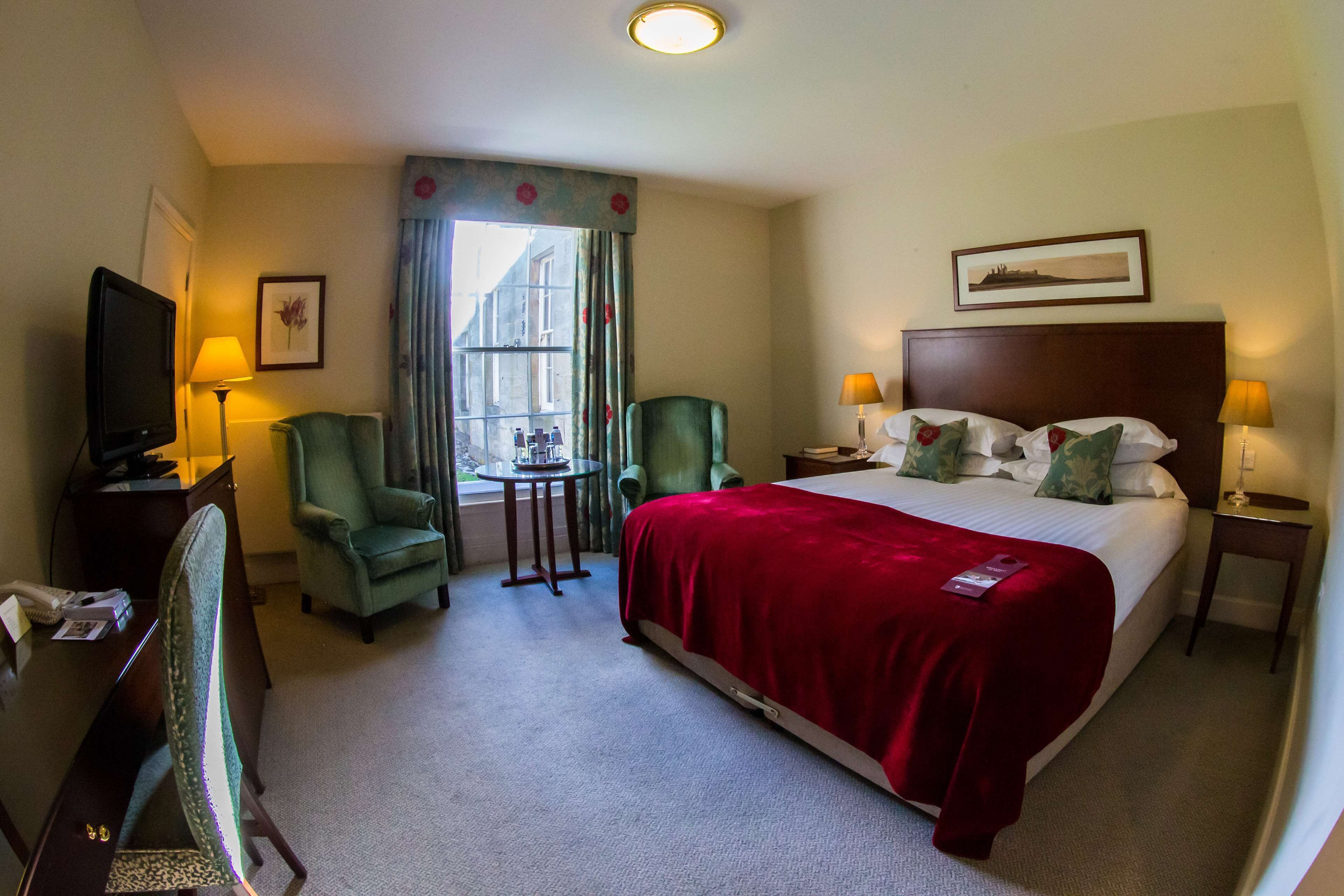 Habitación de hotel con una cama grande, dos sillones, un escritorio con televisor, una mesita y una ventana con cortinas.