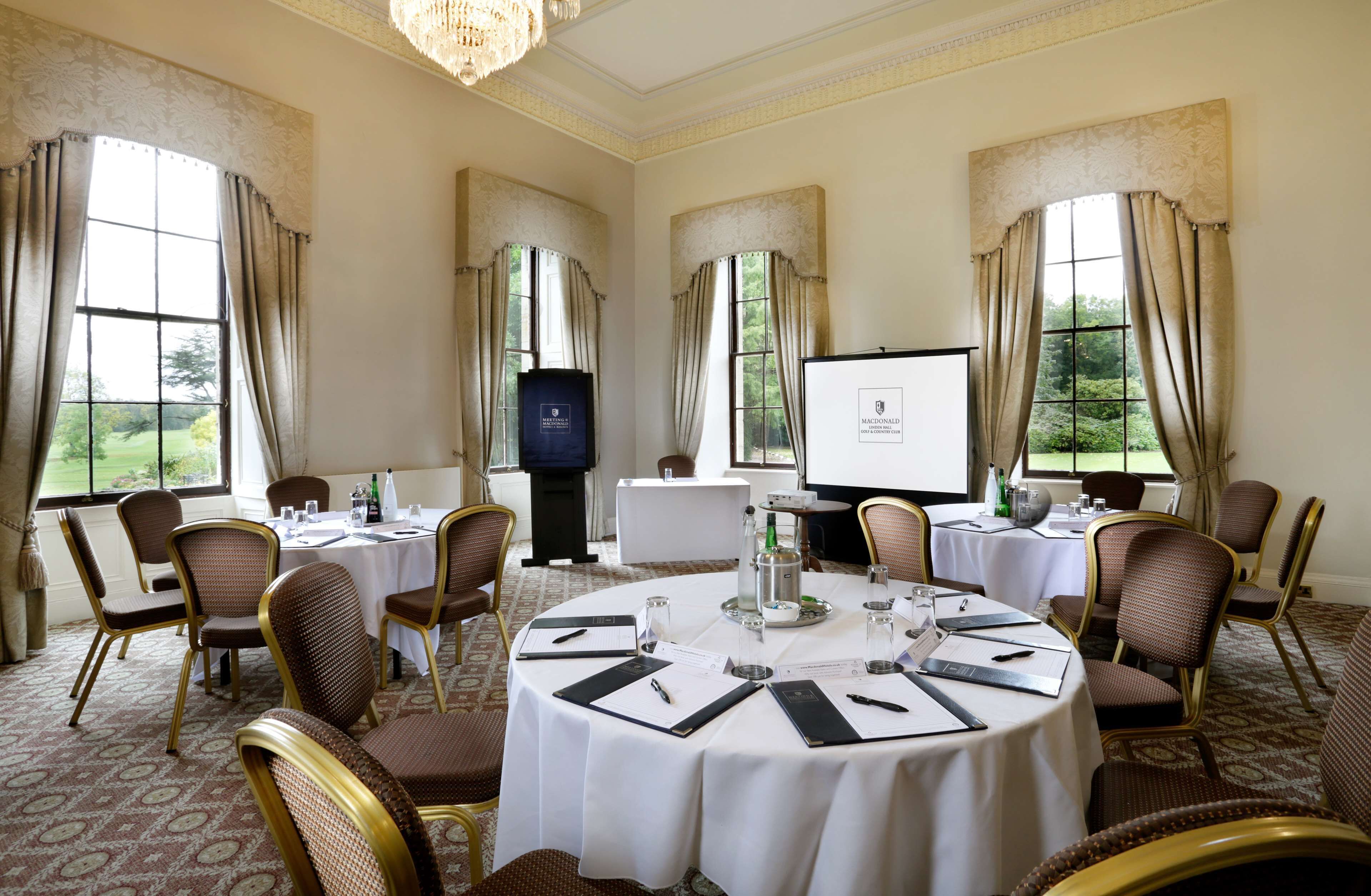 A conference room with round tables, chairs, a projector screen, and a view of greenery outside.