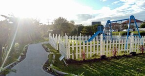 A playground with a white picket fence, a concrete path, and greenery. - Hotel Cueli (Santillana del Mar)