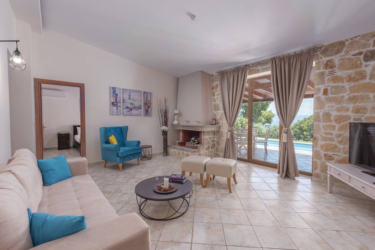 Un salon spacieux avec un canapé beige, un fauteuil bleu et une table basse. On y trouve une cheminée en pierre, un téléviseur et une vue sur une piscine.
