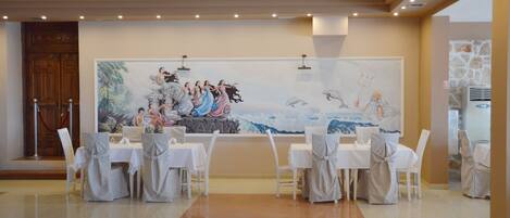 A dining area with white tables and chairs, a large mural on the wall, and a wooden door with a red rope barrier.