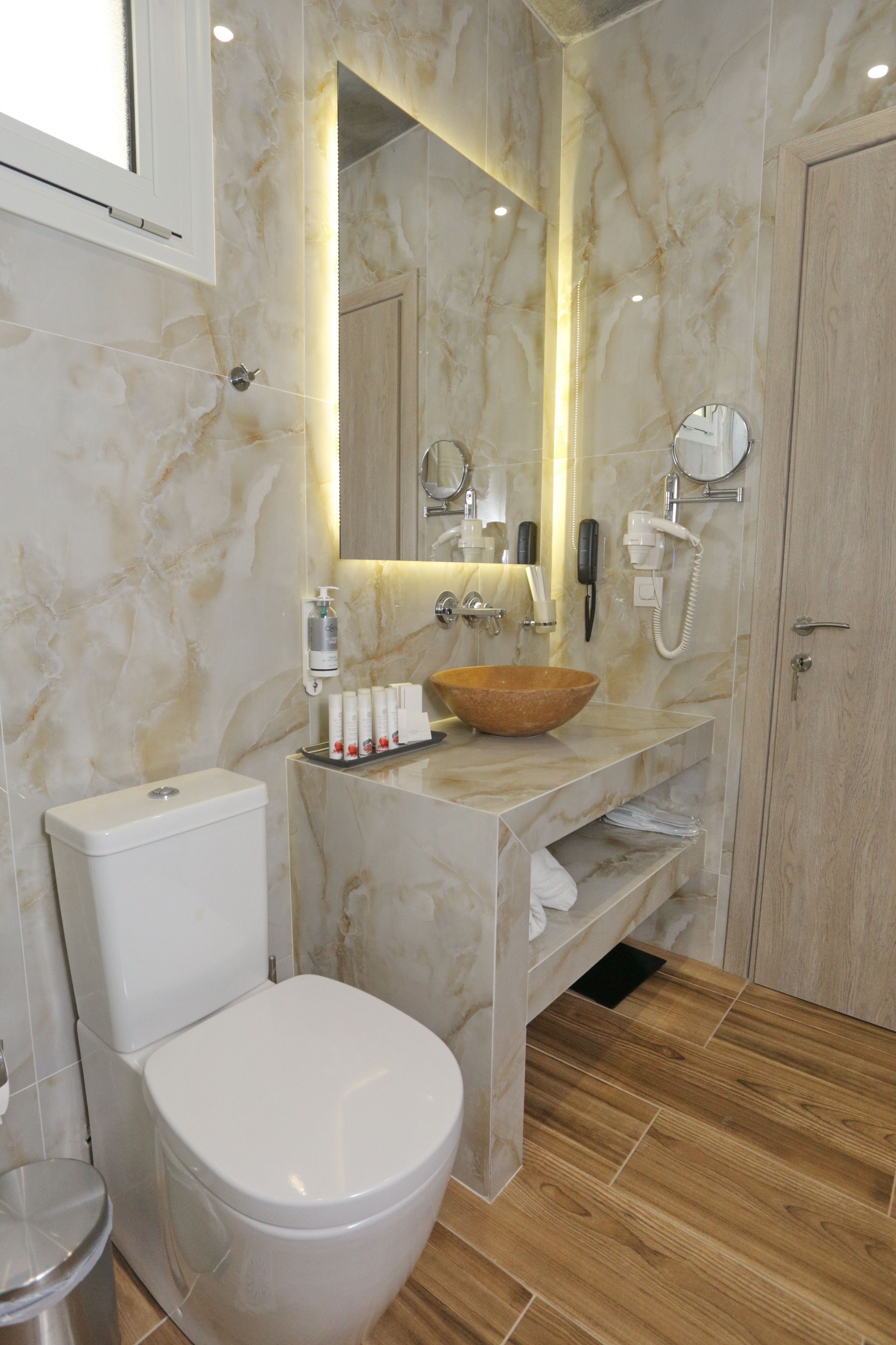 A modern bathroom with a marble vanity, a wooden bowl sink, a toilet, and a mirror with a phone and hairdryer mounted on the wall.