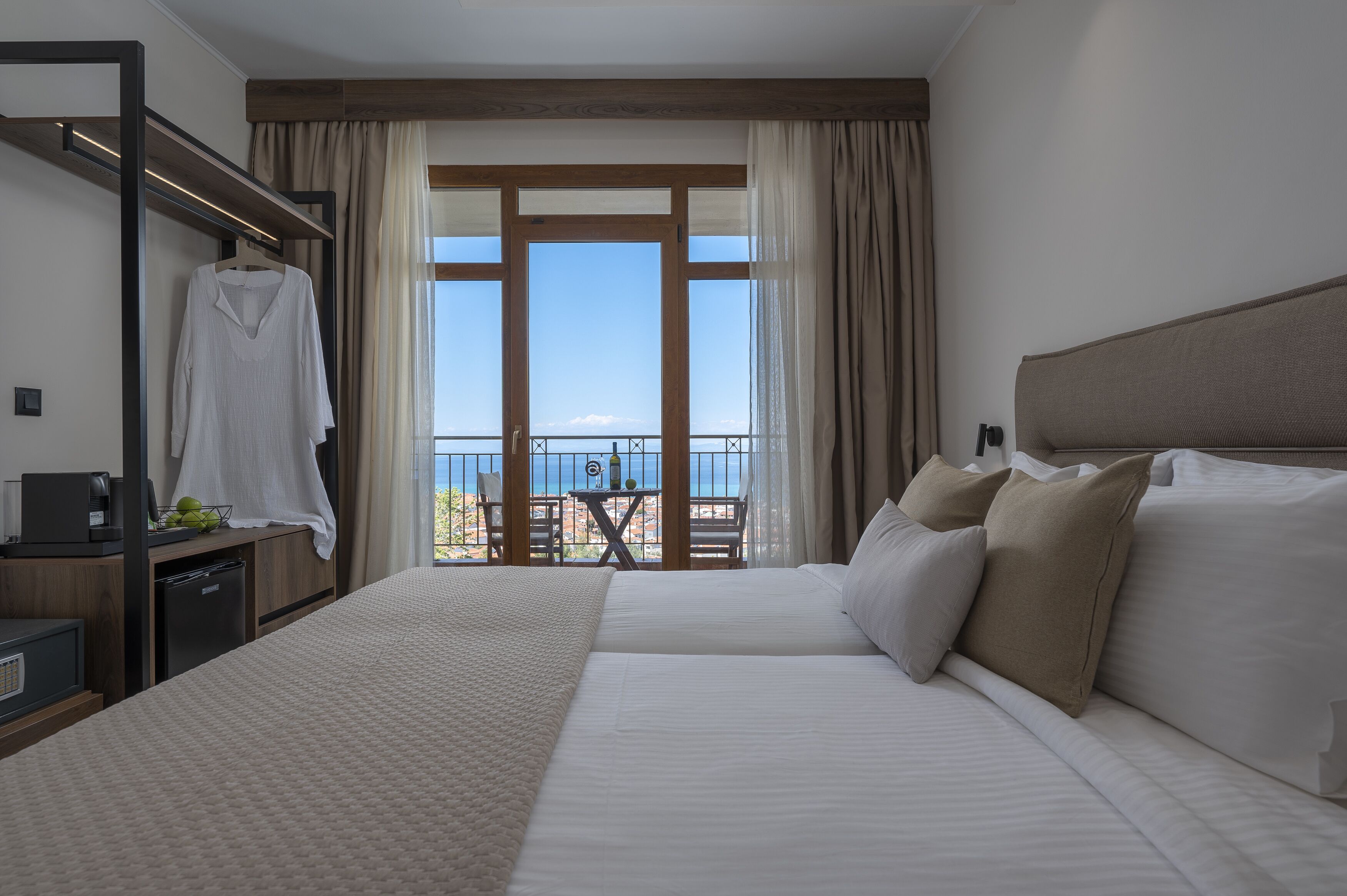 A hotel room with a large bed, a desk with a coffee maker, a hanging robe, and a balcony with a table and chairs.