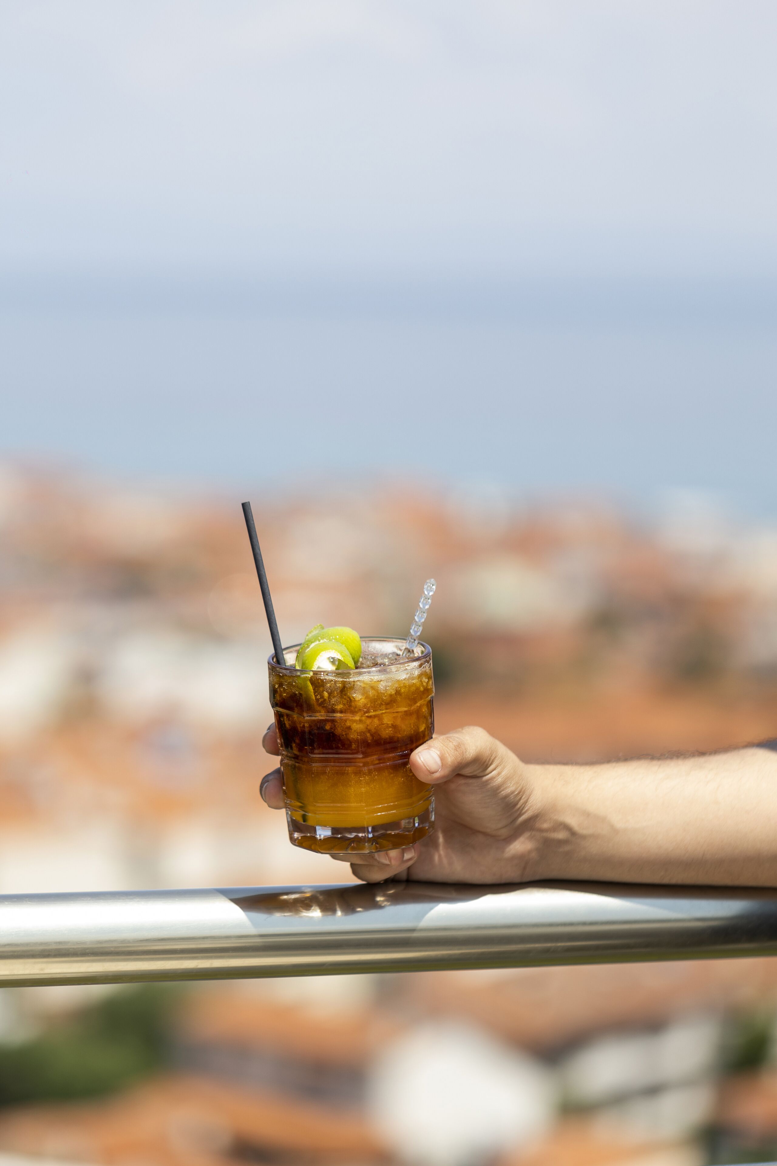 A hand holding a glass of iced tea with a straw and a lime slice, overlooking a cityscape.