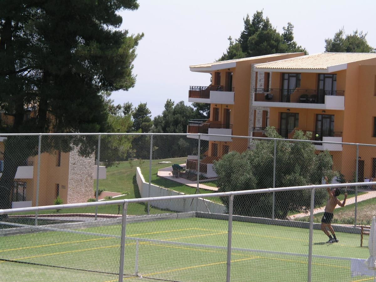 A tennis court with a net, surrounded by a fence. There are buildings in the background.