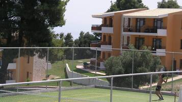 A tennis court with a net, surrounded by a fence. There are buildings in the background.