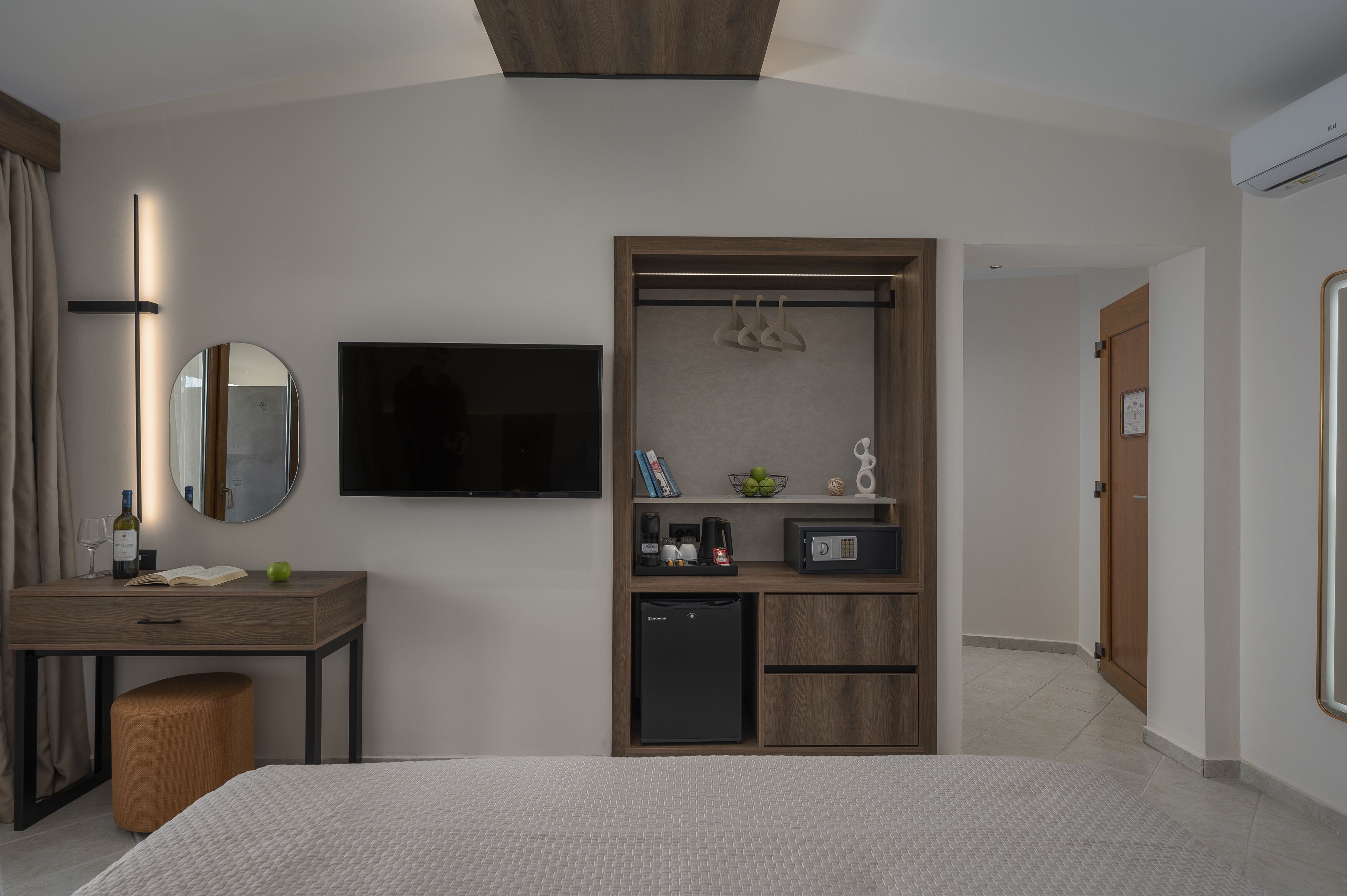 A hotel room with a bed, a desk with a mirror, a television, a minibar, and a wardrobe with a hanging organizer.