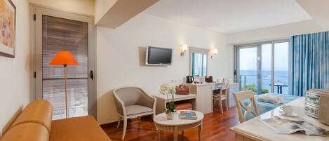 A modern hotel room with a sofa, a dining table, a TV, a desk, and a balcony with a view.