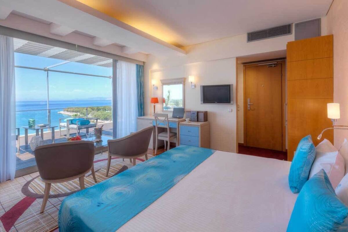 A hotel room with a bed, desk, chairs, a TV, and a balcony with a view of the ocean.
