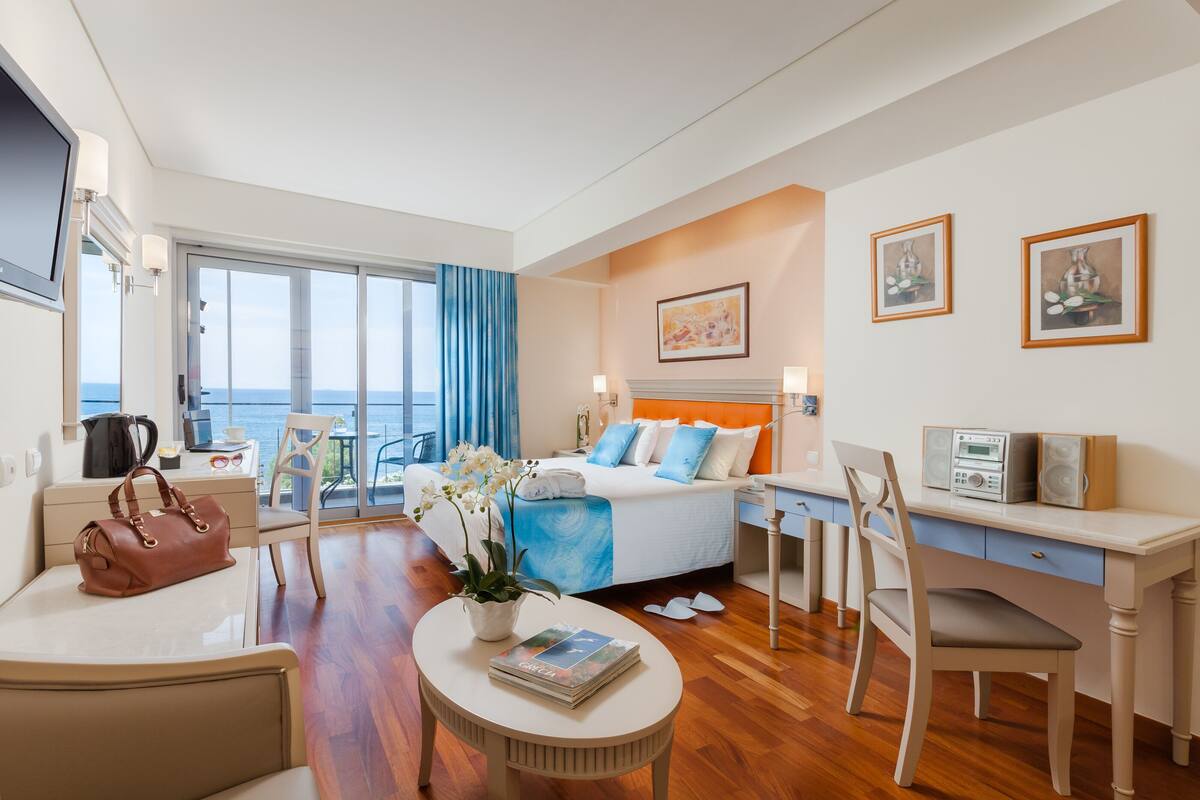 A modern hotel room with a bed, desk, chair, and a view of the sea.