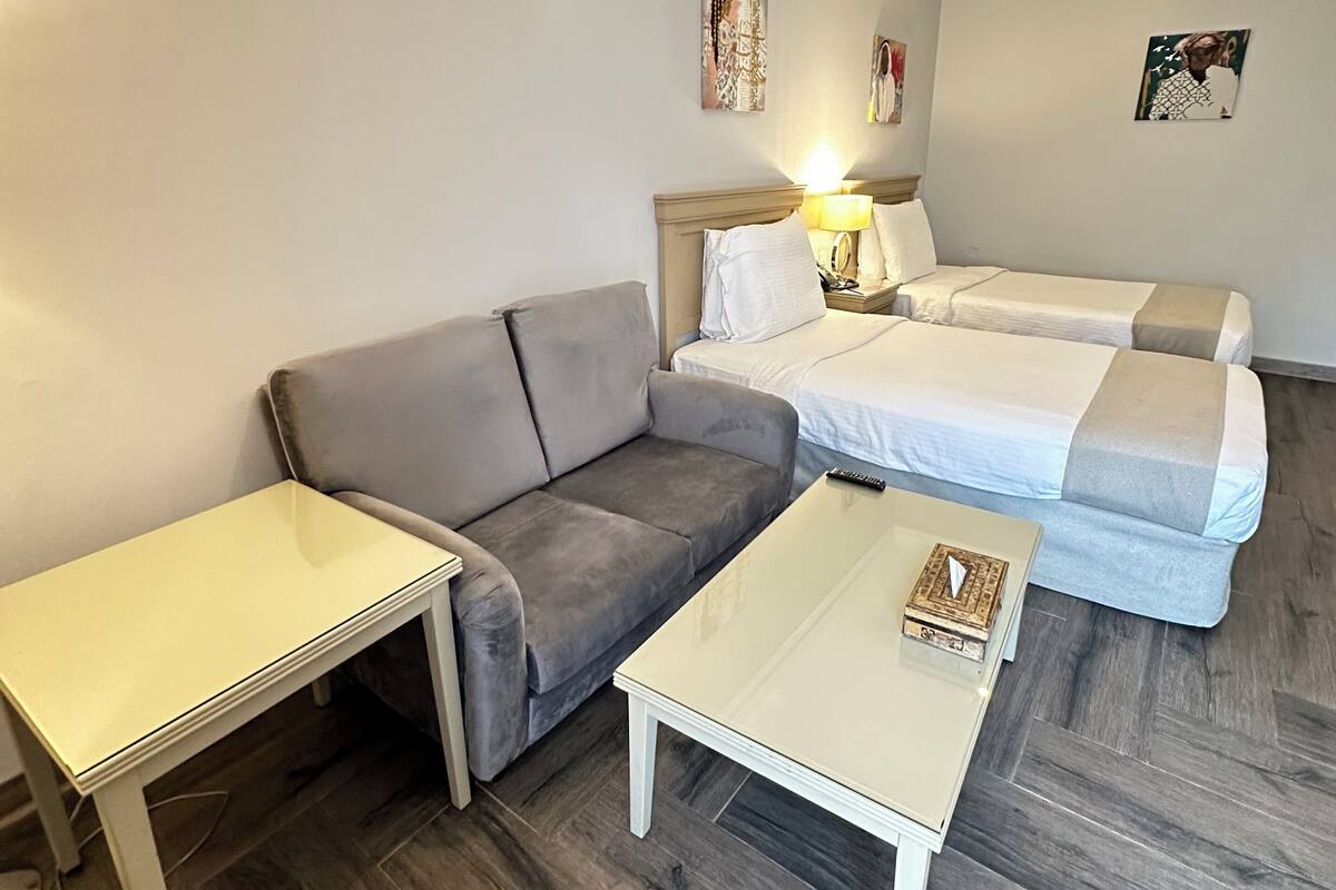 Quarto de hotel compacto com um sofá cinzento, uma pequena mesa branca e uma cama com edredom cinza e branco.